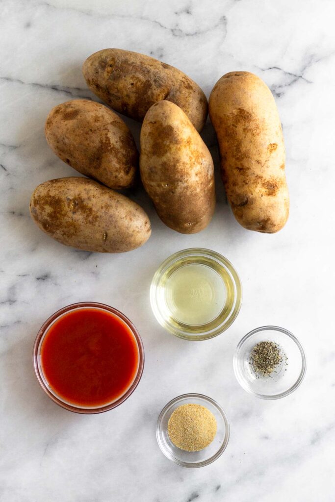 A Mable counter with a handful of russet potatoes, a bowl of oil, a bowl of salt and pepper, a bowl of garlic powder, and a bowl of hot sauce.