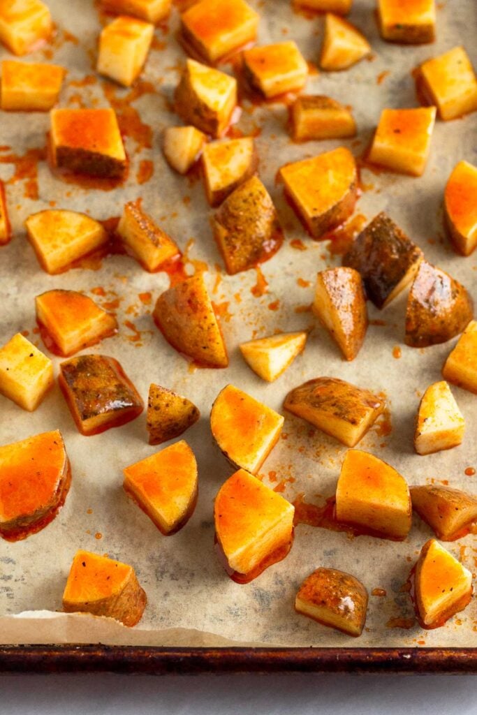 Baking sheet lined with parchment paper with diced potatoes coated in buffalo sauce spread out on it.