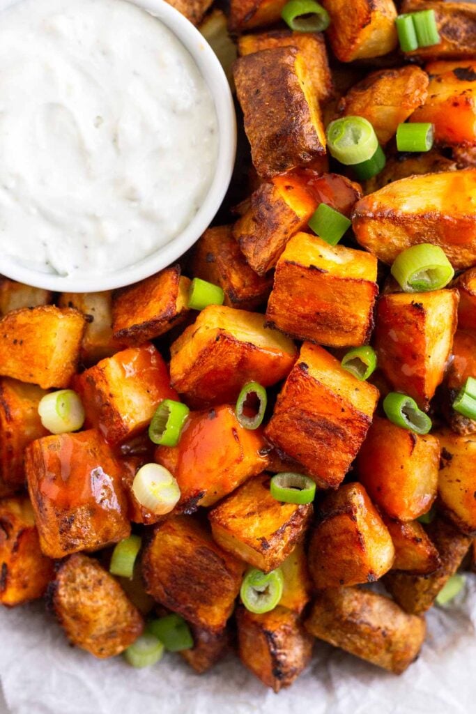 Close up of buffalo potatoes on a plate with a ramekin of blue cheese dressing next to them. They are topped with hot sauce and green onions.