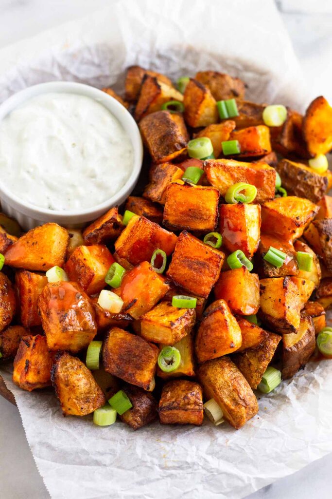 Buffalo potatoes topped with hot sauce and green onions on a plate with a ramekin of blue cheese dressing next to them.