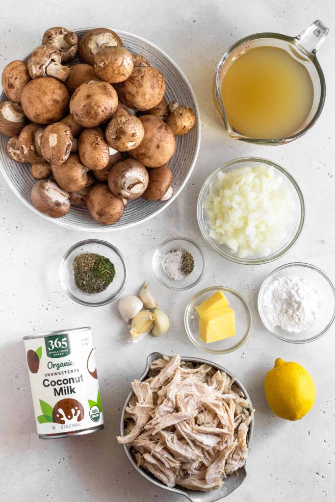 White counter with a bowl of mushrooms, a measuring cup of broth, a bowl of diced onions, a bowl of flour, a whole lemon, a bowl of shredded chicken, a can of full-fat coconut milk, 4 cloves of garlic, a bowl of salt and pepper, and a bowl of spices.