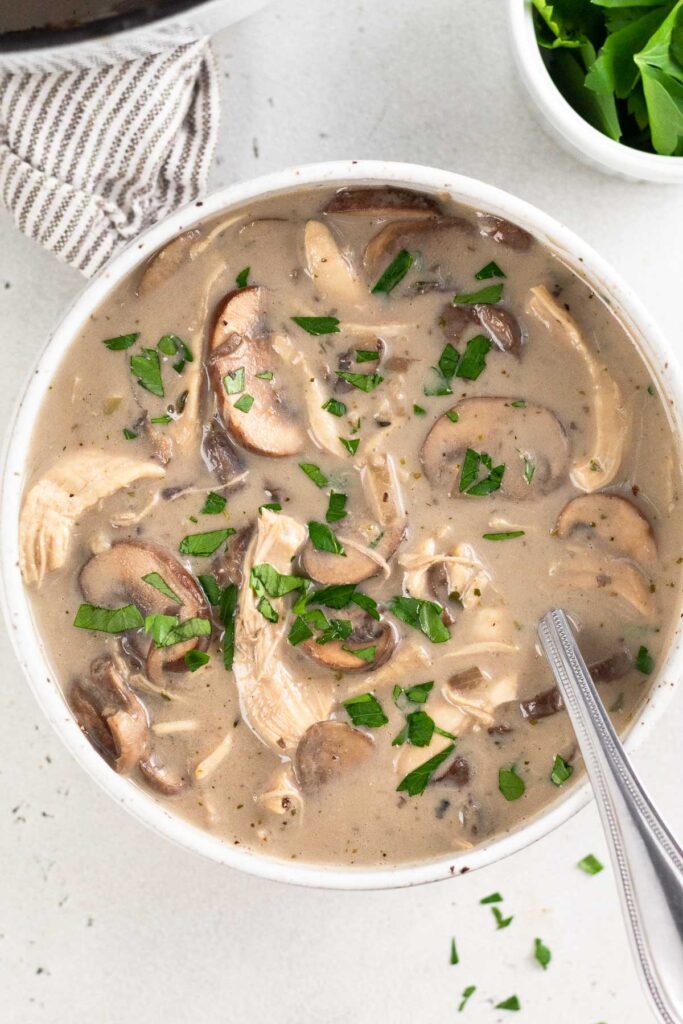 Overhead shot of a bowl of chicken and mushroom soup with a spoon in it and topped with fresh parsley. Next to it is a dish of more parsley and a kitchen towel.