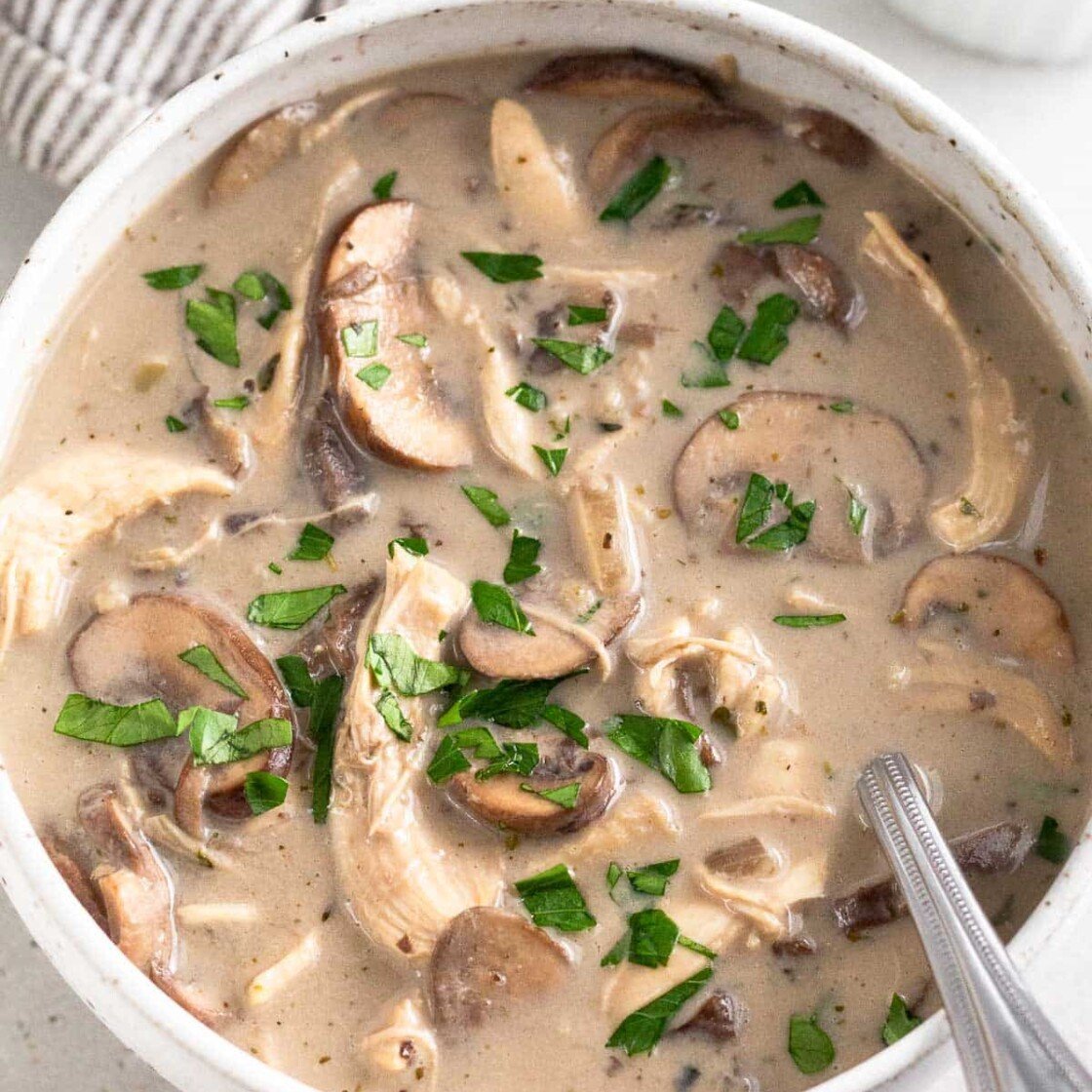 A bowl of creamy chicken mushroom soup topped with fresh parsley. A spoon is in the bowl. Behind it is a ramekin of more herbs and big pot.