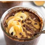Chocolate peanut butter overnight oats in a jar topped with sliced banana, peanut butter, and cacao nibs with a spoon in the jar. Behind it is a spoon with peanut butter on it.