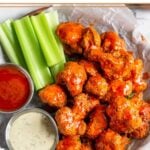 Buffalo cauliflower on a plate with a side of celery, a small dish of buffalo sauce, and a small dish of ranch dressing.