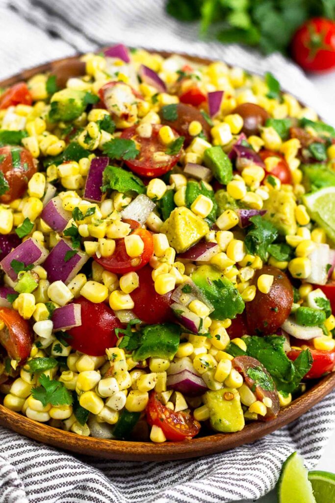 Corn salsa with tomatoes, avocado, cilantro, and jalapeños in a dish. Around it are some lime wedges, cilantro, and cherry tomatoes.