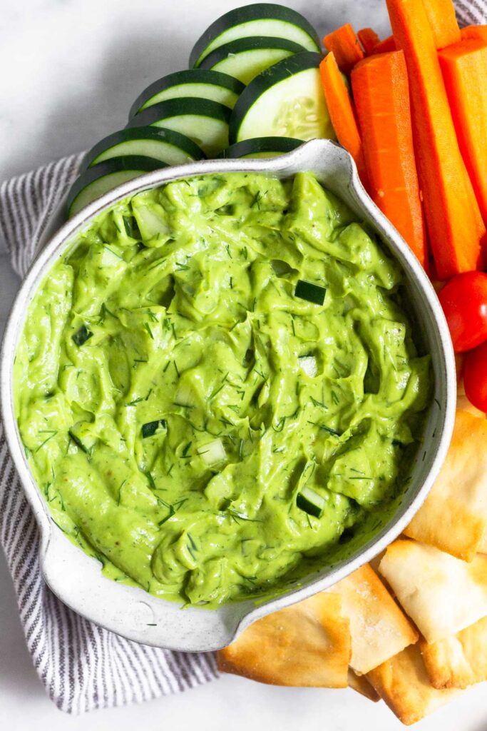 Avocado tzatziki in a bowl with veggies and pita chips around it.