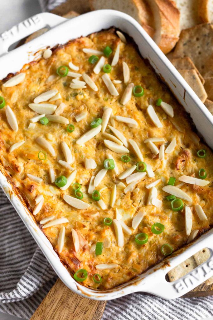 A baking dish filled with crab dip topped with silvered almonds green onions. Around it is some bread and crackers.