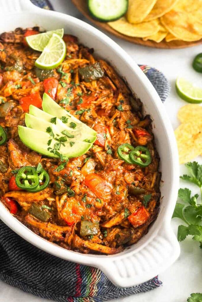 Healthy super bowl recipe of chicken enchilada dip in a baking dish topped with avocado, cilantro, jalapeños, and lime wedges. Behind it is a plate of chips.