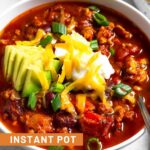 A bowl of turkey chili topped with avocado, sour cream, shredded cheese, and green onions with a spoon in the bowl. Behind it a bowl of tortilla chips and ramekin of sliced green onions.