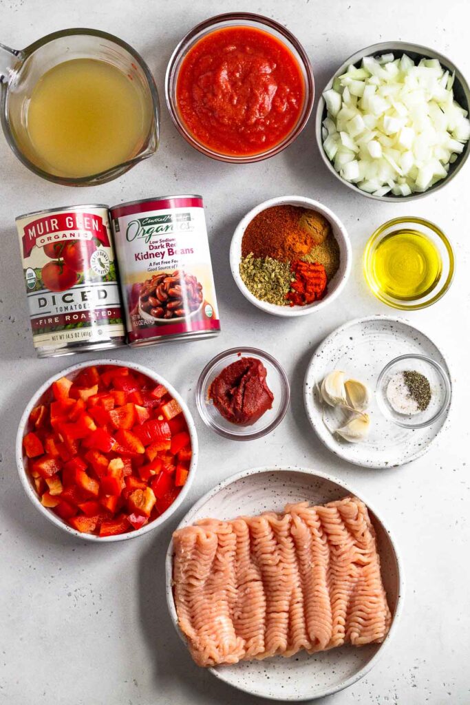 White counter with bowl of tomato sauce, a bowl of diced onions, a small bowl of olive oil, a bowl of spices, a plate of garlic cloves and a small bowl of salt and pepper, a plate of raw ground turkey, a bowl of diced bell peppers, a bowl of tomato paste, a can of kidney beans, a can of diced tomatoes, and a cup of broth.