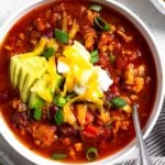Instant pot turkey chili in a white bowl topped with sour cream, avocado, shredded cheese, and green onions. There is also a spoon in the bowl. Around the bowl is a plate of sliced jalapeños, a dish of sliced green onions, a bowl of tortilla chips, and a striped kitchen towel.