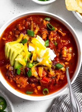 Instant pot turkey chili in a white bowl topped with sour cream, avocado, shredded cheese, and green onions. There is also a spoon in the bowl. Around the bowl is a plate of sliced jalapeños, a dish of sliced green onions, a bowl of tortilla chips, and a striped kitchen towel.