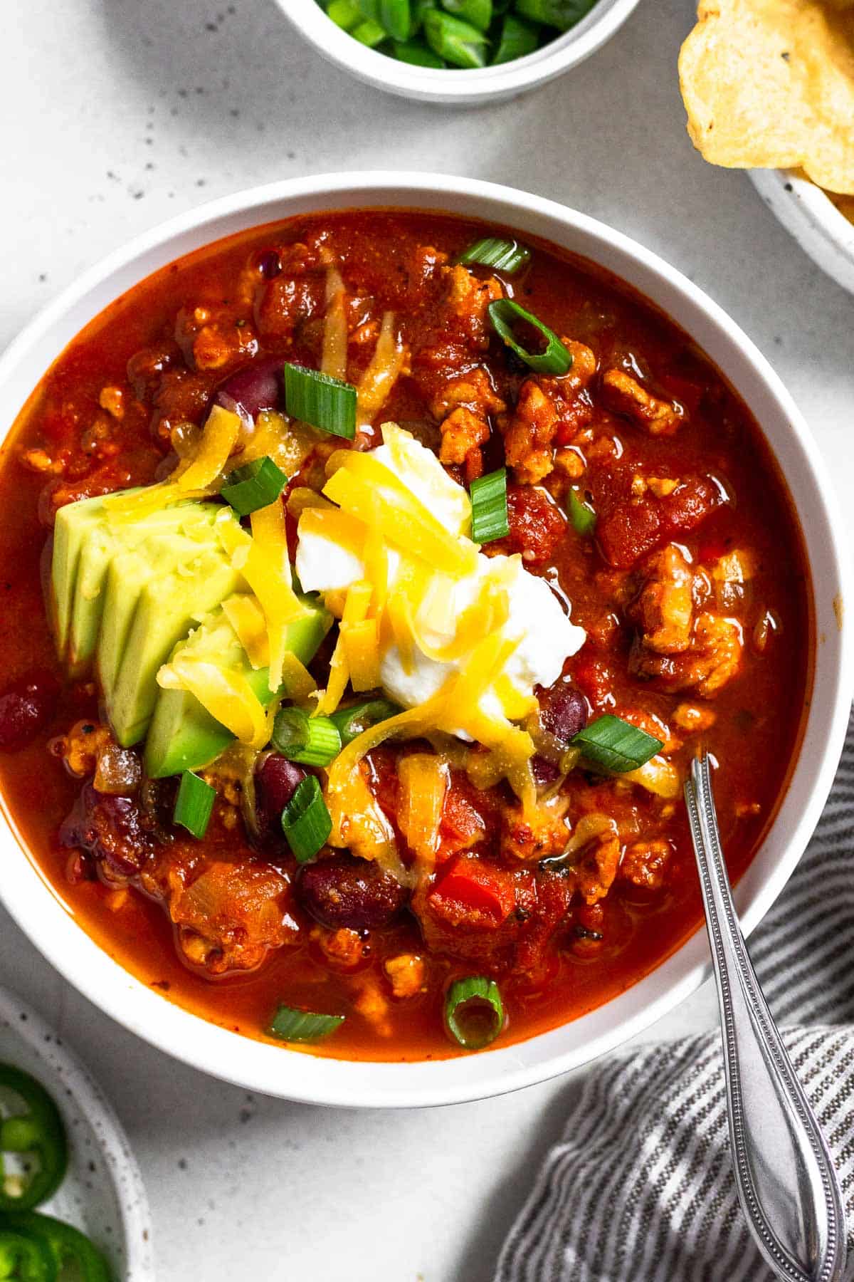 Instant pot turkey chili in a white bowl topped with sour cream, avocado, shredded cheese, and green onions. There is also a spoon in the bowl. Around the bowl is a plate of sliced jalapeños, a dish of sliced green onions, a bowl of tortilla chips, and a striped kitchen towel.
