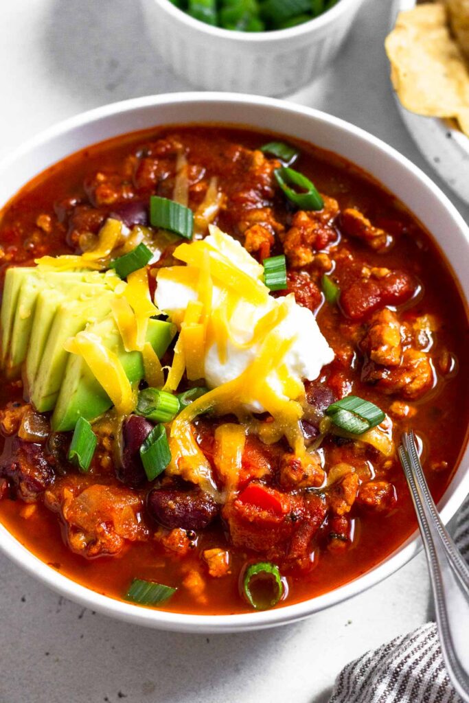 A close up of a bowl of instant pot turkey chili topped with avocado, sour cream, shredded cheese, and green onions. A spoon is also in the bowl. Behind the bowl is a bowl of tortilla chips and ramekin of sliced green onions.