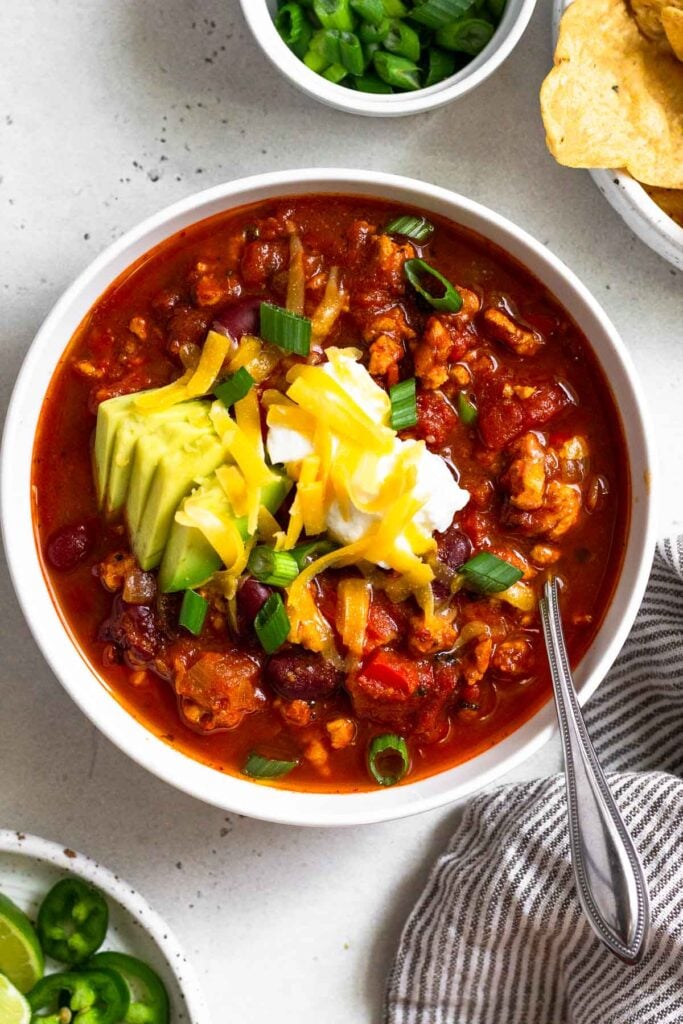 Healthy turkey chili in a white bowl topped with sour cream, avocado, shredded cheese, and green onions. There is also a spoon in the bowl. Around the bowl is a plate of sliced jalapeños, a bowl of sliced green onions, a bowl of tortilla chips, and a striped kitchen towel.