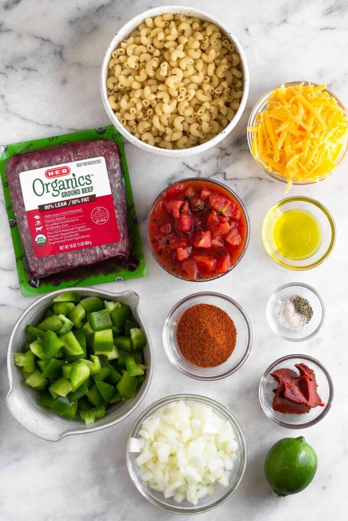 A bowl of elbow macaroni, a bowl of shredded cheddar cheese, a bowl of canned tomatoes, a small bowl of olive oil, a small bowl of salt and pepper, a bowl of tomato paste, a lime, a bowl of diced onions, a small bowl of spices, a bowl of diced green bell peppers, and a package of raw ground beef on a white marble counter.
