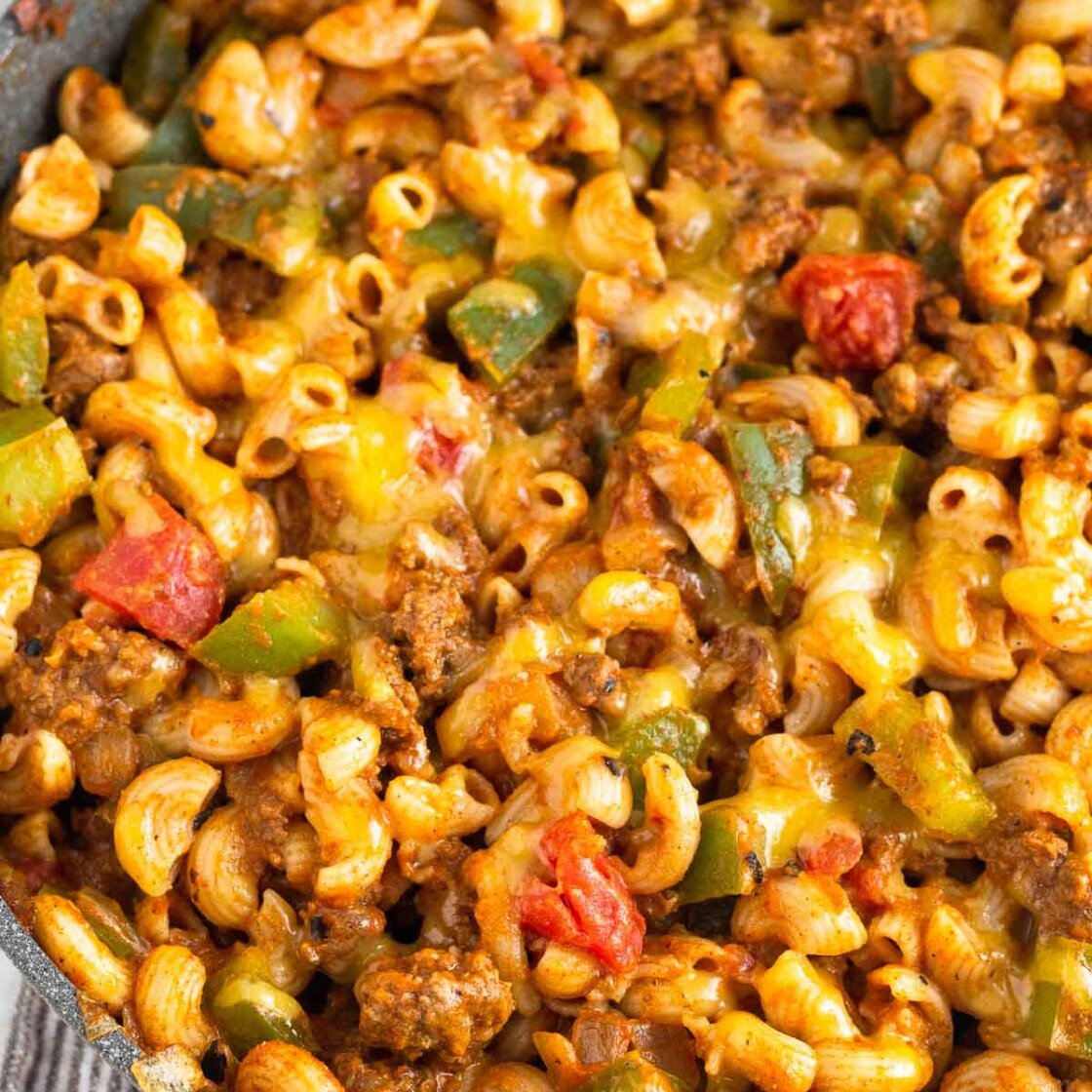 Cheesy taco pasta with ground beef and veggies in a skillet. The skillet is on a kitchen towel and there is a ramekin of fresh herbs behind it.
