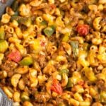 Cheesy taco pasta with ground beef and veggies in a skillet. The skillet is on a kitchen towel and there is a ramekin of fresh herbs behind it.