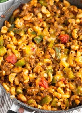 Cheesy taco pasta with ground beef and veggies in a skillet. The skillet is on a kitchen towel and there is a ramekin of fresh herbs behind it.