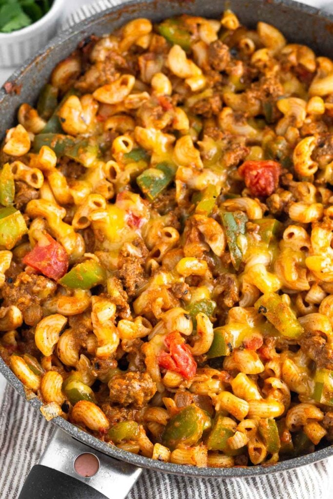 Cheesy taco pasta with ground beef and veggies in a skillet. The skillet is on a kitchen towel and there is a ramekin of fresh herbs behind it.