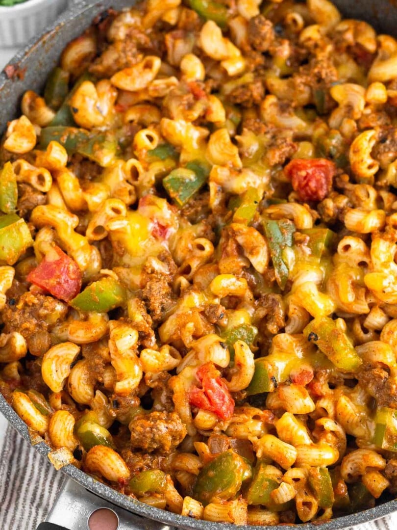 Cheesy taco pasta with ground beef and veggies in a skillet. The skillet is on a kitchen towel and there is a ramekin of fresh herbs behind it.