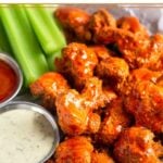 A plate of crispy air fryer buffalo cauliflower covered in buffalo sauce. Also on the plate is cut up celery, a ramekin of ranch, and a ramekin of buffalo sauce.