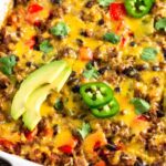 Close up of cheesy beef burrito casserole in a large casserole dish. It is topped with sliced avocado, sliced jalapeño, and cilantro.