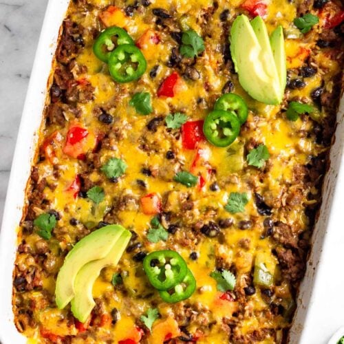 Burrito casserole in a large casserole dish garnished with sliced avocado, sliced jalapeño, and cilantro. Next to it is half an avocado and ramekin of cilantro.