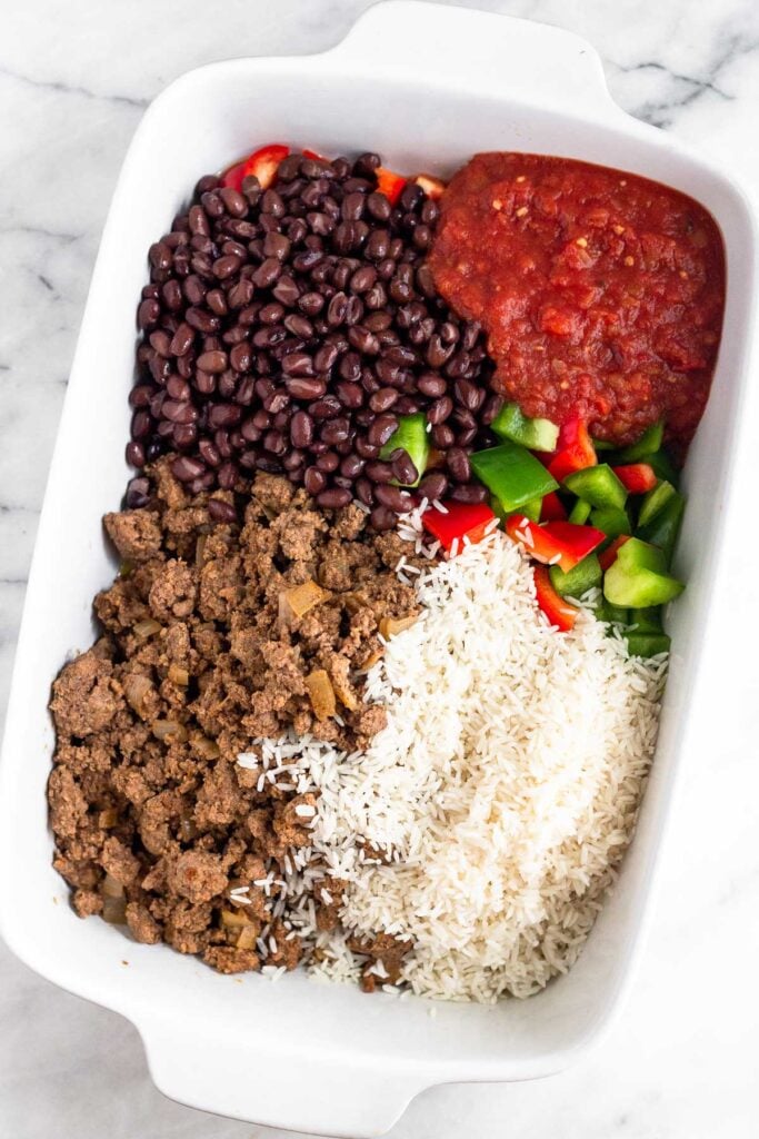 Large casserole dish filled with seasoned ground beef, black beans, salsa, diced bell peppers, and uncooked white rice. Each thing is in individual piles next to each other.