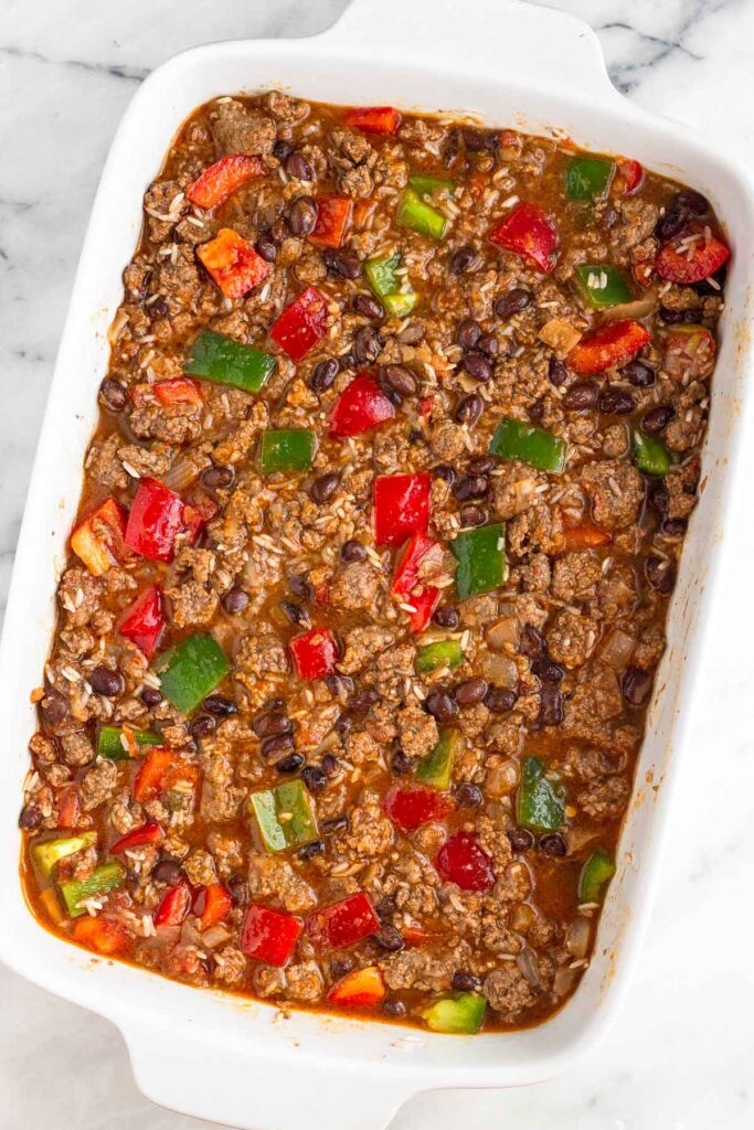A large casserole dish filled with a mixture of ground beef, rice, beans, diced bell peppers, and broth all mixed together. The broth is just covering the ingredients.