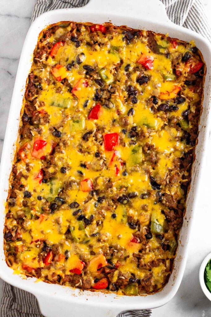 Cheesy burrito casserole in large casserole dish. The dish is on top of a dish towel with a small bowl of herbs to the side.