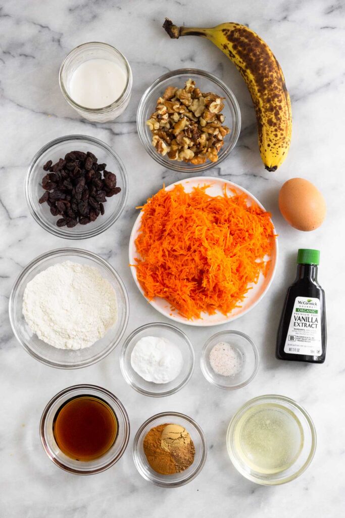 White marble counter with a brown banana, an egg, a bottle of vanilla extract, a bowl of coconut oil, a bowl of spices, a bowl of maple syrup, a bowl of salt, a bowl of baking powder and baking soda, a bowl of flour, a plate of shredded carrots, a bowl of raisins, a bowl of walnuts, and a jar of milk.