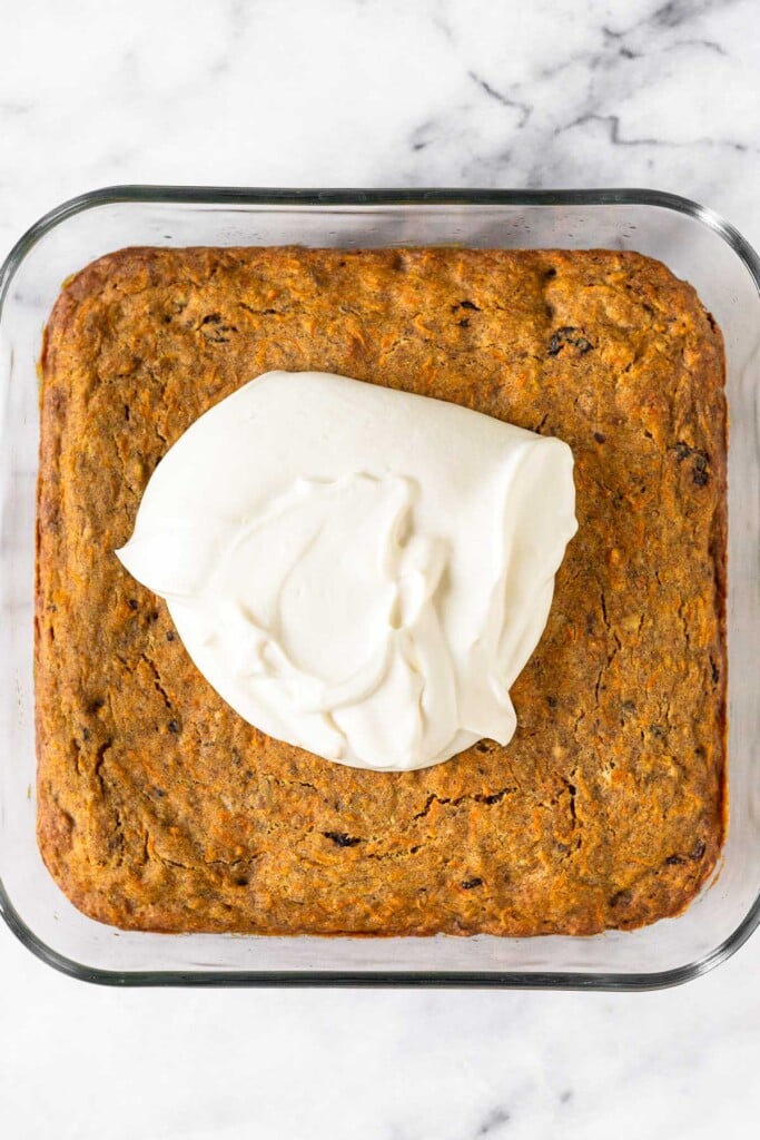 Carrot cake in a glass baking dish with a mound of greek yogurt icing on top of them before it is spread.