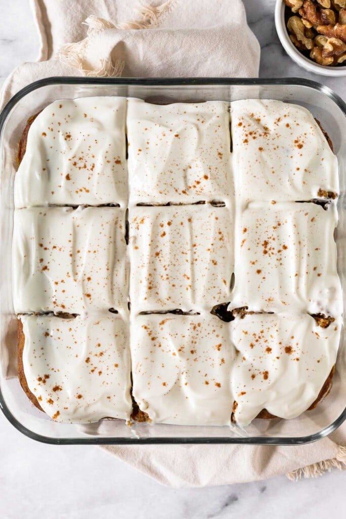 Carrot cake bars topped with frosting and sprinkled with cinnamon in a glass baking dish. They are cut into 9 equal-sized pieces. The dish is on a white linen with a dish of walnuts next to it.
