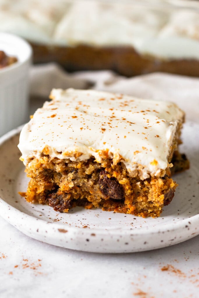 A piece of healthy carrot cake topped with frosting and sprinkled with cinnamon on a small plate. Behind it is a pan with the rest of the cake.