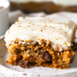 Carrot cake bar topped with frosting and cinnamon on a small plate. Behind it is the pan of the rest of the bars.