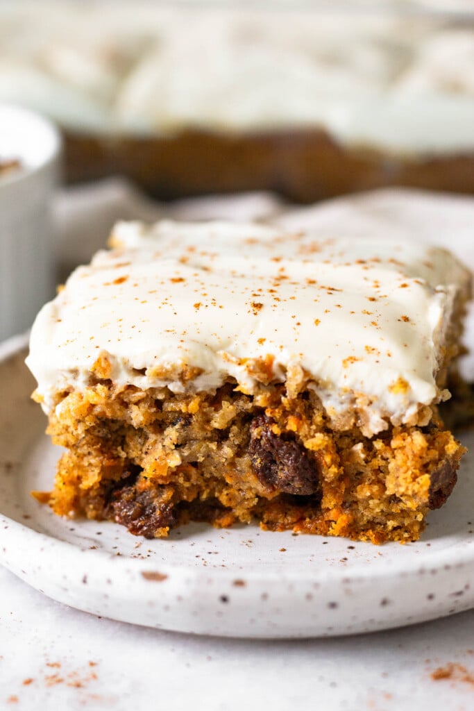 Carrot cake bar topped with frosting and cinnamon on a small plate. Behind it is the pan of the rest of the bars.