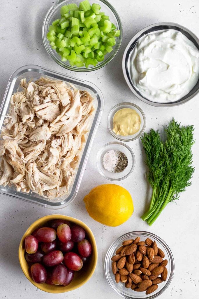 White concrete counter with a bowl of diced celery, a bowl of greek yogurt, a few sprigs of fresh dill, a small bowl of dijon mustard, a small bowl of salt and pepper, a whole lemon, a bowl of almonds, a bowl of red grapes, and a glass container of shredded chicken.