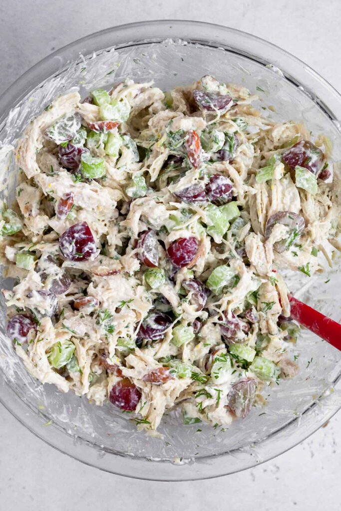 Chicken salad with grapes, almonds, celery, and dill in a large glass bowl with a spatula in it.