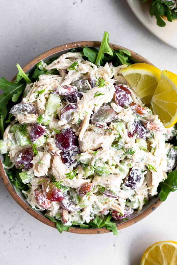 Healthy chicken salad with grapes and almonds in a bowl with arugula underneath it. It is garnished with 2 lemon wedges. Half a lemon is next to the bowl.