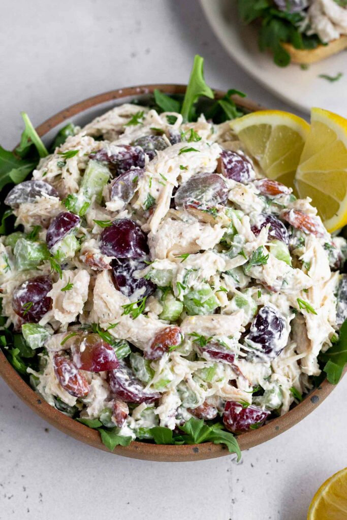 Greek yogurt chicken salad with grapes and almonds in a bowl overtop some arugula garnished with two lemon wedges. There is half a lemon next to the plate and a plate of more chicken salad in the background.