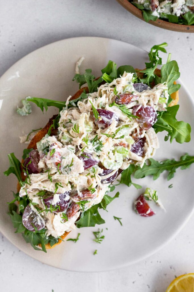 Overhead shot of chicken salad with greek yogurt, grapes, and almonds on top of some arugula on a piece of toast on a plate.