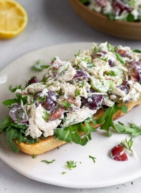 Healthy chicken salad on top of some arugula on a piece of toast on a white plate. Behind it is half a lemon and a bowl of more chicken salad.