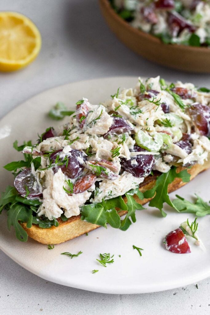 Healthy chicken salad on top of some arugula on a piece of toast on a white plate. Behind it is half a lemon and a bowl of more chicken salad.