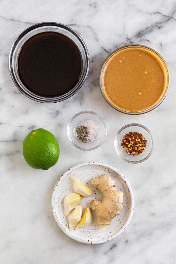 A white marble counter top with a bowl of coconut aminos, a bowl of peanut butter, a bowl of red pepper flakes, a plate of fresh ginger and 4 garlic cloves, a lime, and a small bowl of salt and pepper.