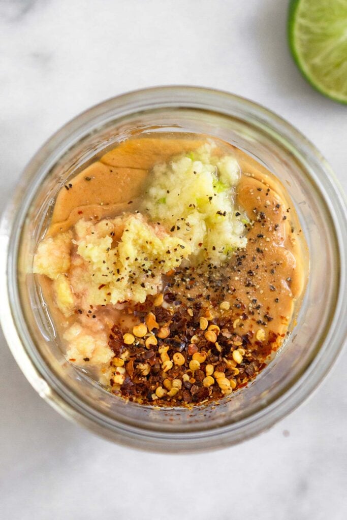 An overhead shot of a jar with peanut butter, coconut aminos, garlic, ginger, lime juice, and red pepper flakes in it. Next to it an half a lime.
