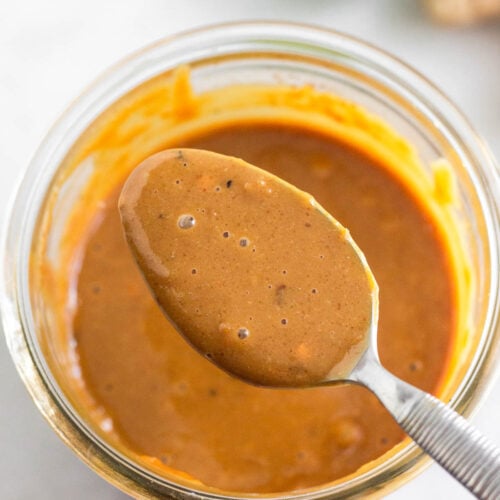 Peanut sauce in a jar with a spoon overtop holding some. Behind the jar is half a lime and fresh ginger.