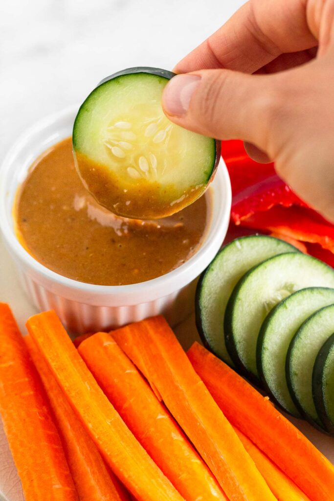A plate of sliced carrots, cucumbers, and bell peppers with a ramekin of peanut butter sauce on it. There is a hand dipping a cucumber into it.
