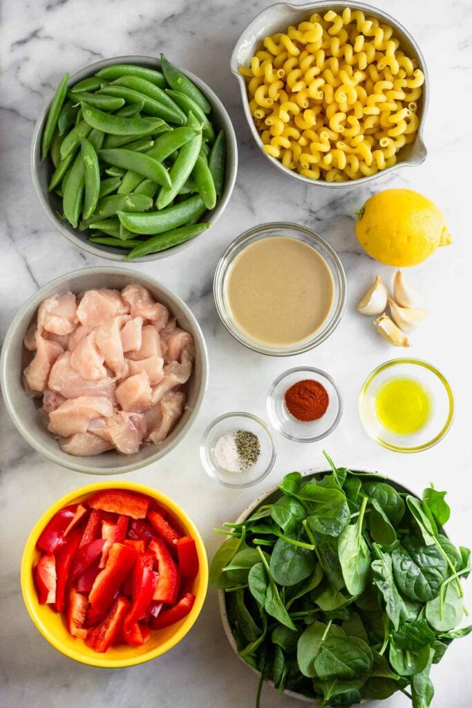 Marble counter top with a bowl of uncooked pasta, a whole lemon, four cloves of garlic, a bowl of oil, a bowl of spinach, a bowl of sliced bell peppers, a bowl of diced raw chicken, two small bowls of spices, a bowl of tahini, and a bowl of snap peas.